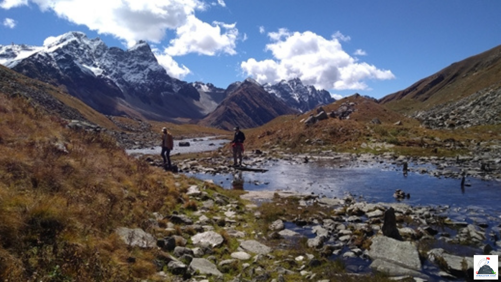 Two trekkers by a shallow stream in a high alpine meadow below jagged, snow-dusted peaks