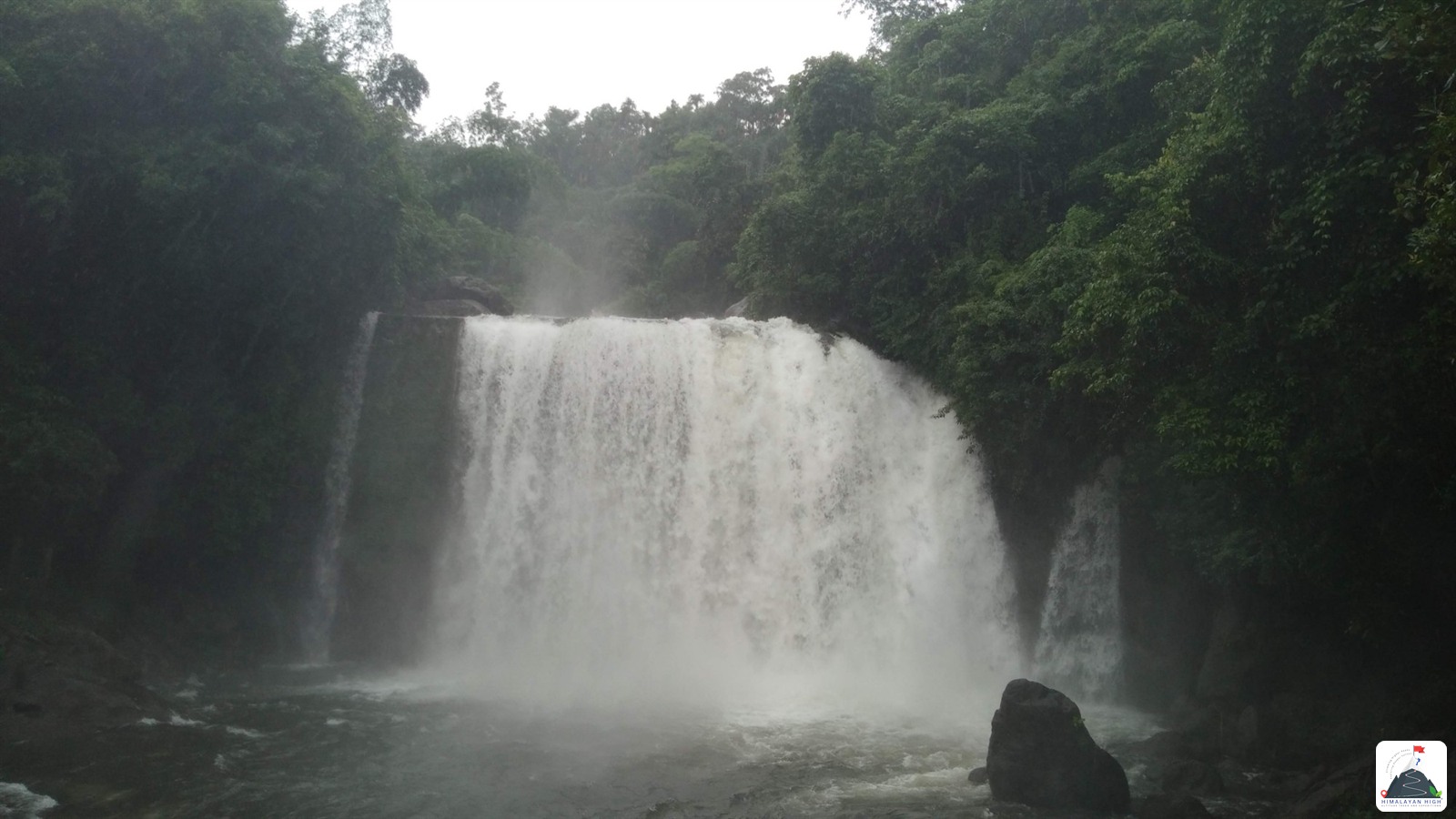 A broad waterfall pouring into a churning pool, framed by dense green forest under a white sky